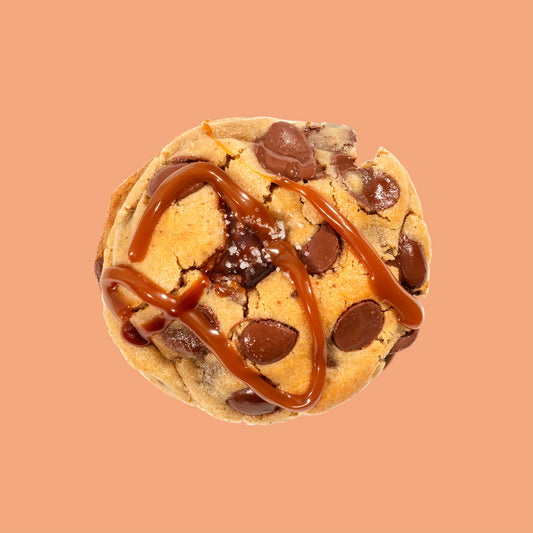 Cookie artisanal au caramel beurre salé et fleur de sel vu de dessus, décoré d'un filet de caramel et garni de grosses pépites de chocolat au lait Callebaut sur fond beige orangé chaud