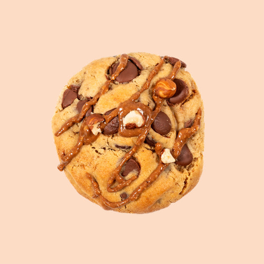 Cookie artisanal au praliné vu de dessus, garni de pépites de chocolat au lait Callebaut, de noisettes entières torréfiées, décoré d'un filet de praliné fondant sur fond clair