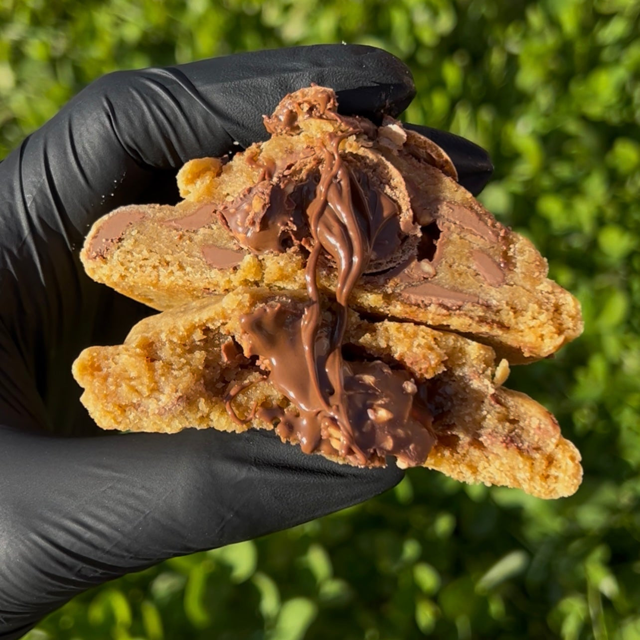 Cookie artisanal au Ferrero rocher coupé en deux, tenu en main avec un gant noir, révélant un cœur fondant et gourmand de Ferrero Rocher, chocolat noisette, éclats de noisettes torréfiées et pépites de chocolat au lait Callebaut. Texture moelleuse, visuel généreux et ultra gourmand sur un fond végétal vert
