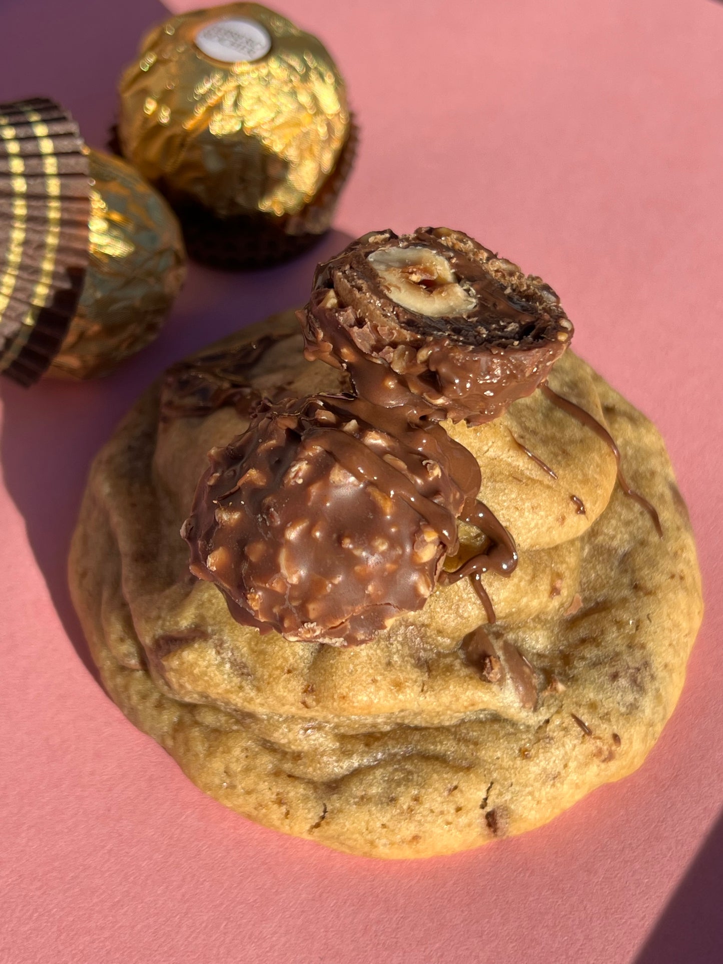Cookie artisanal Ferrero Rocher, présenté sur fond rose, garni d'un chocolat Ferrero coupé en deux laissant apparaître son cœur fondant et sa noisette entière. Texture moelleuse, chocolat fondant et éclats de noisettes visibles. En arrière-plan, des Ferrero Rocher entiers apportent une touche premium et gourmande.  