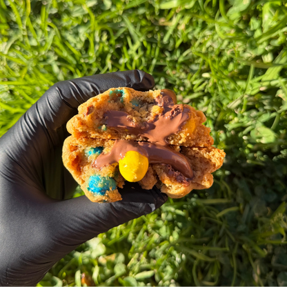 Cookie M&M's artisanal coupé en deux, avec cœur fondant de Nutella coulant, tenu à la main, révélant une pâte vanillée moelleuse et des M&M's croquants, sur fond végétal vert.  