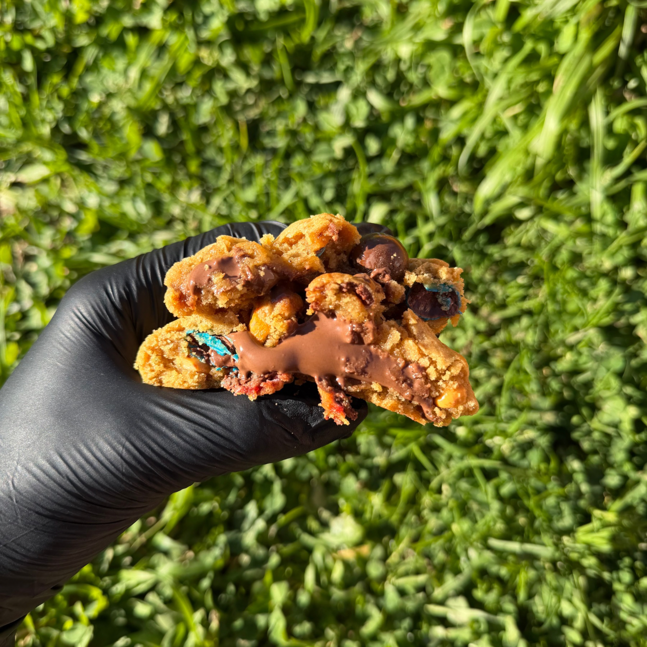 Cookie M&M’s artisanal tenu à la main, avec cœur fondant de Nutella coulant, pâte vanillée moelleuse et M&M’s croquants visibles, sur fond végétal vert en extérieur.