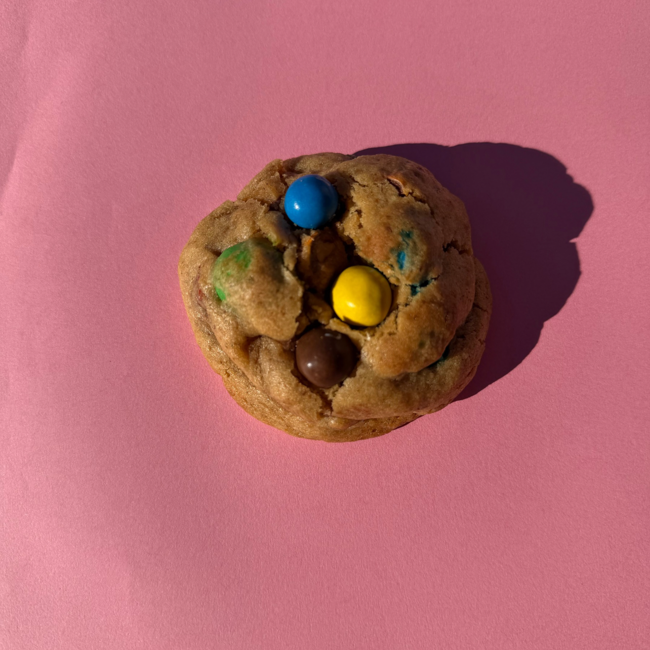 Cookie M&M’s artisanal vu du dessus, pâte vanillée moelleuse et légèrement dorée, garni de M&M’s colorés croquants, présenté sur un fond rose.