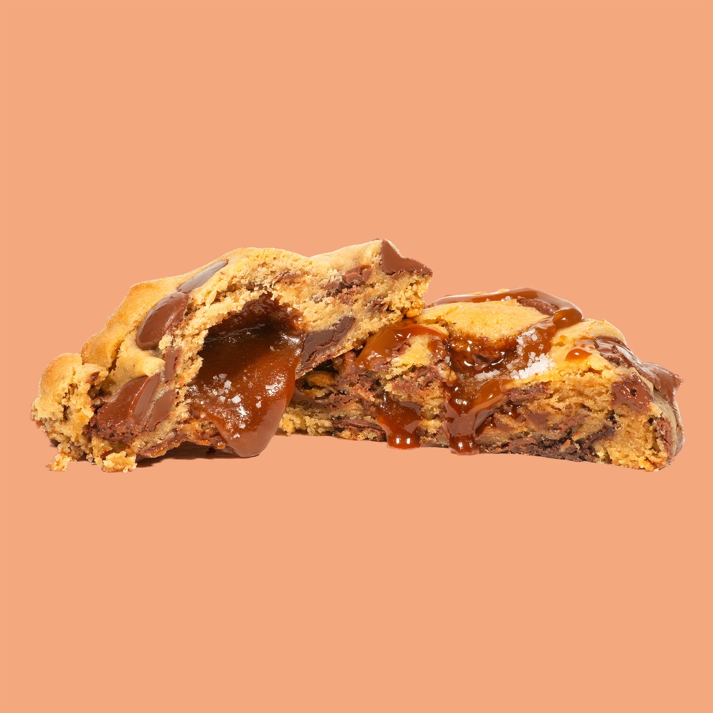 Cookie artisanal au caramel beurre salé et fleur de sel, coupé en deux pour révéler un cœur coulant de caramel doré et de généreuse pépites de chocolat au lait Callebaut. Texture fondante et moelleuse sur fond beige orangé chaud 