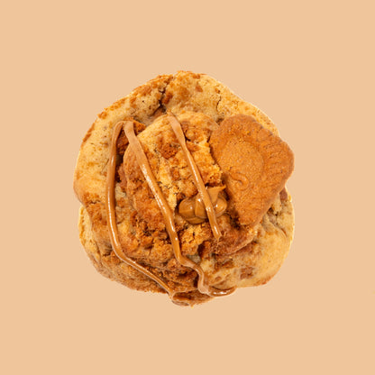 Cookie artisanal au spéculos vu de dessus, décoré d'un filet de pâte de spéculos lotus et d'un biscuit et brisures de spéculos croustillant sur fond beige orangé chaud. Texture moelleuse et dorée