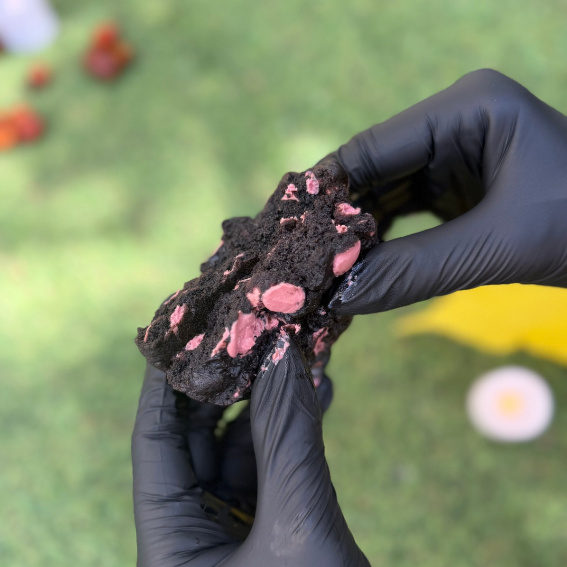 Cookie artisanal Halloween ouvert en 2 au cacao noir Callebaut garni de chocolat ruby Callebaut, tenu avec 2 mains, avec une texture moelleuse et fondante sur fond végétal 